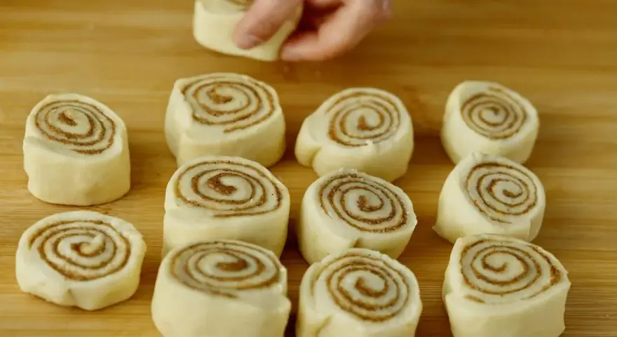 Färska kanelbullar, eller "rollo de canela", ligger redo att bakas på en träskärbräda. En hand placerar en bullform.