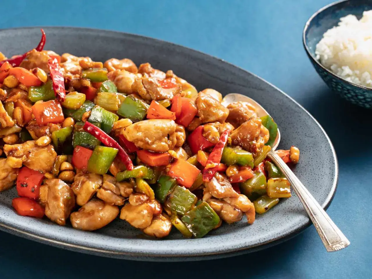 En tallrik med kyckling kung pao, full av färgglada grönsaker, jordnötter och torkad chili. En skål med ris syns i bakgrunden.