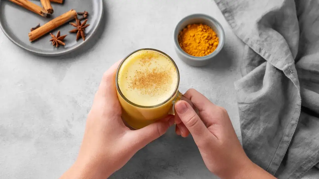 Händer håller ett glas gyllene mjölk med kanel och stjärnanis. Bredvid finns en skål med gurkmejapulver, som visar para q sirve la curcuma.