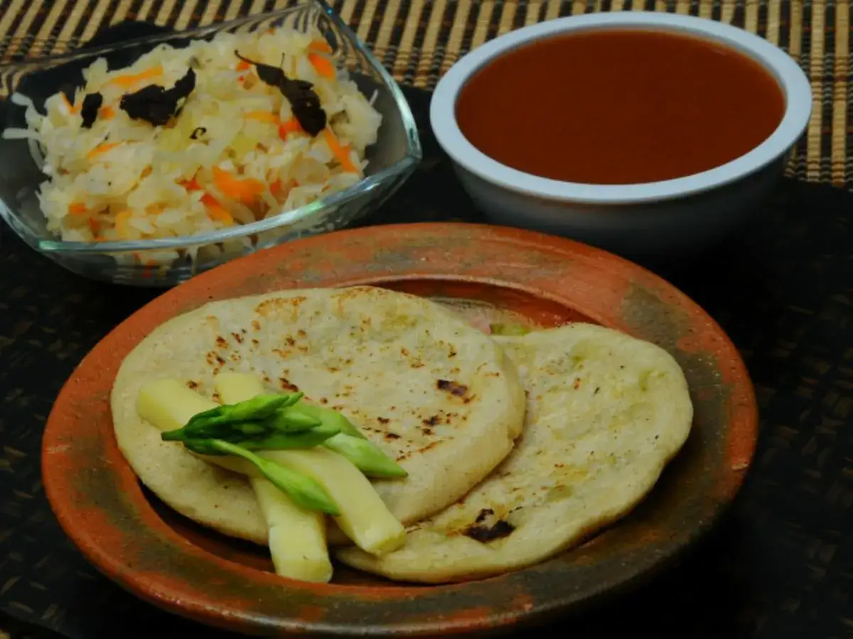 Tv&aring; pupusas de queso con loroco serveras med en sk&aring;l med k&aring;lsallad och en sk&aring;l med r&ouml;d s&aring;s.