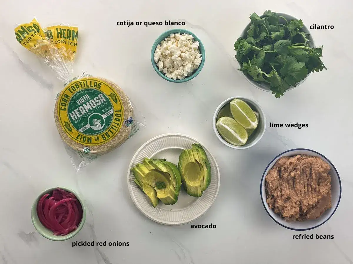 Här är ingredienser för tacos: majstortillas, refried beans, avokado, picklad rödlök, koriander, limeklyftor och cotijaost.