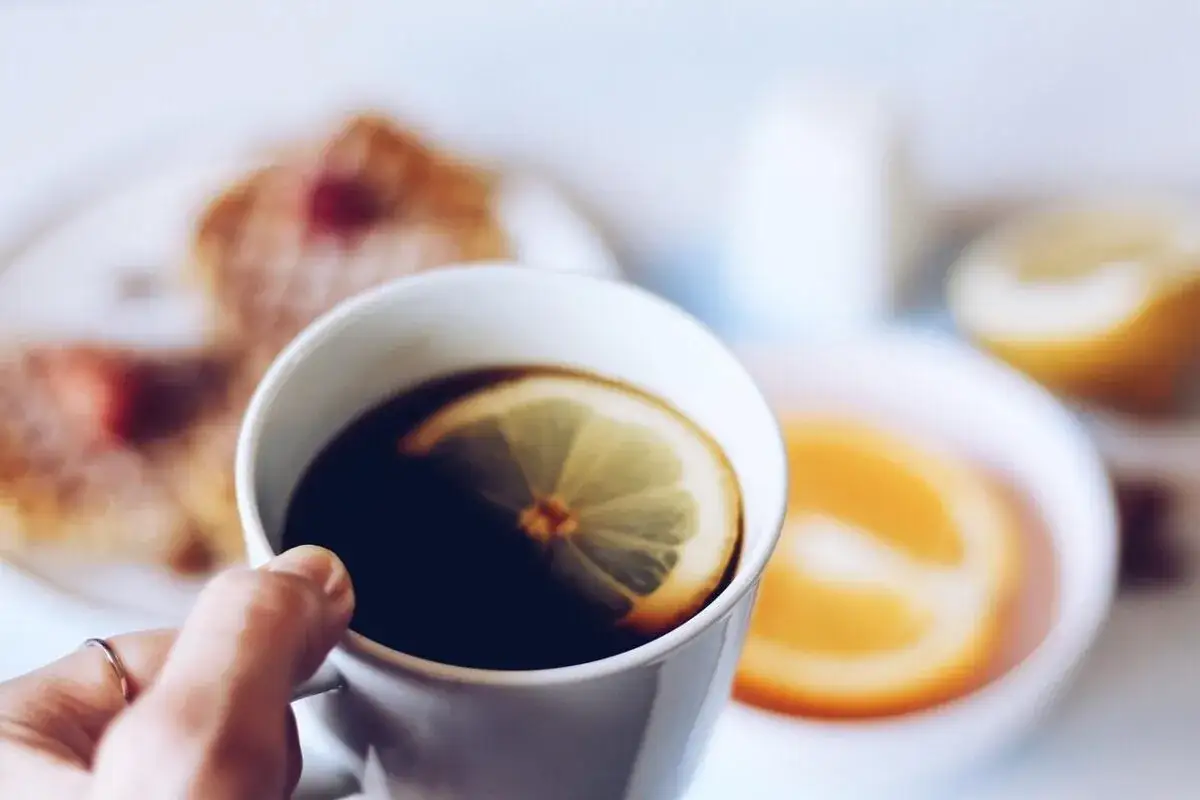 En hand håller en kopp kaffe med en citronskiva, en perfekt cafe con limon för en lugn stund. I bakgrunden syns pannkakor och apelsin.