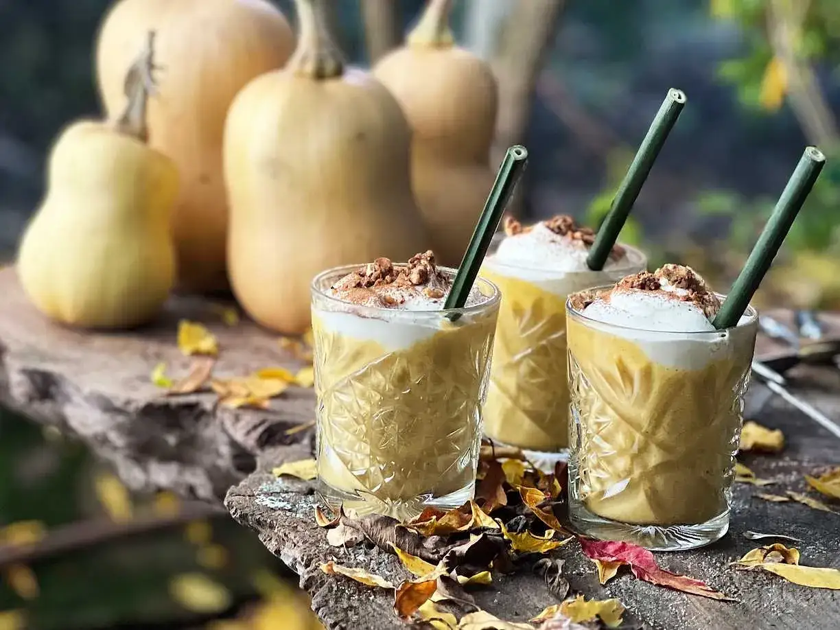 Tre glas med krämig pumpa- och honungsdrink, toppad med grädde och nötter. Pumpan i bakgrunden skvallrar om höst.