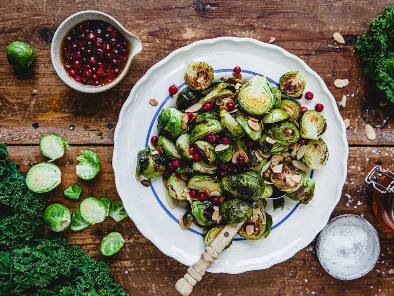 Rostade coles de bruselas med tranbär och mandel, serverade på en vit tallrik med blå kant. En skål med dressing och färsk grönkål ligger bredvid.