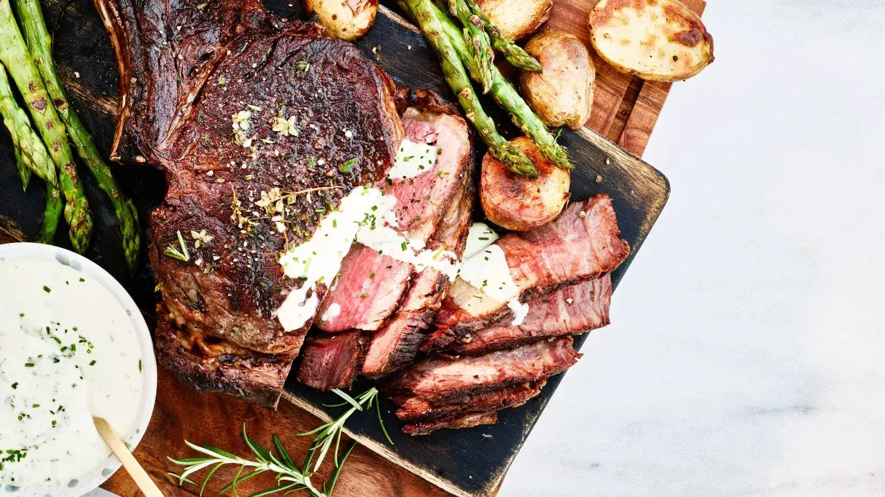 En saftig tomahawk steak skuren i skivor, serverad med rostade potatisar, sparris och en krämig sås.