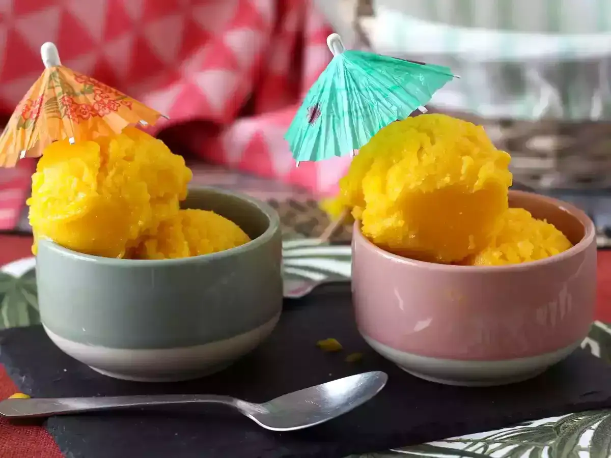 Tv&aring; sk&aring;lar med solgul mango en miel-glass, dekorerade med sm&aring; paraplyer. En sked ligger bredvid.