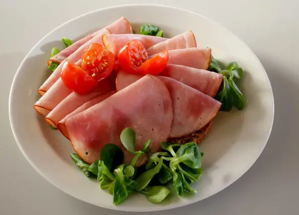 Skivor av saftig skinka, en köttprodukt, serveras med körsbärstomater och gröna blad på en tallrik.