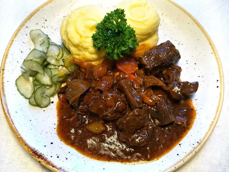 En tallrik med mustigt estofado de res, krämigt potatismos och skivad gurka. En grön persiljekvist toppar moset.